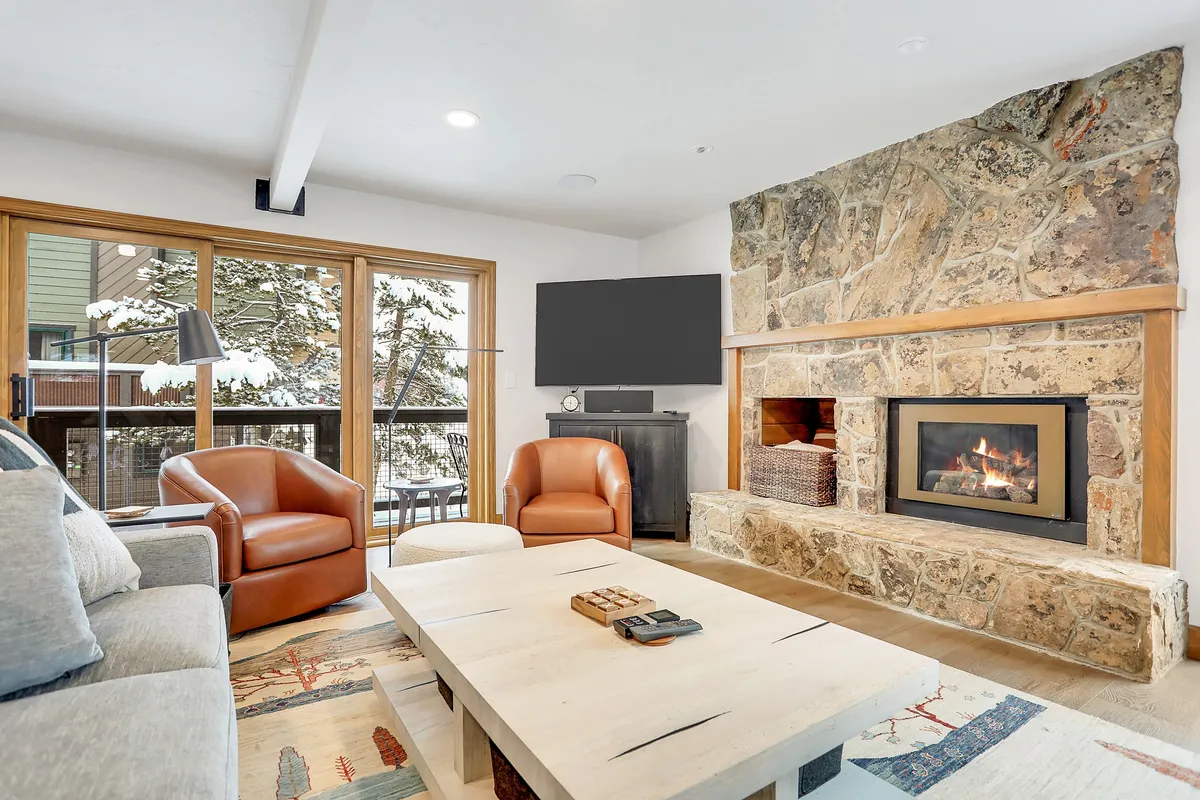 A cozy living space featuring a modern fireplace, expansive windows framing snowy mountain views, comfortable seating, and a coffee table. The space invites relaxation, socializing, and enjoying the warmth and ambiance of the fire after a day outside.