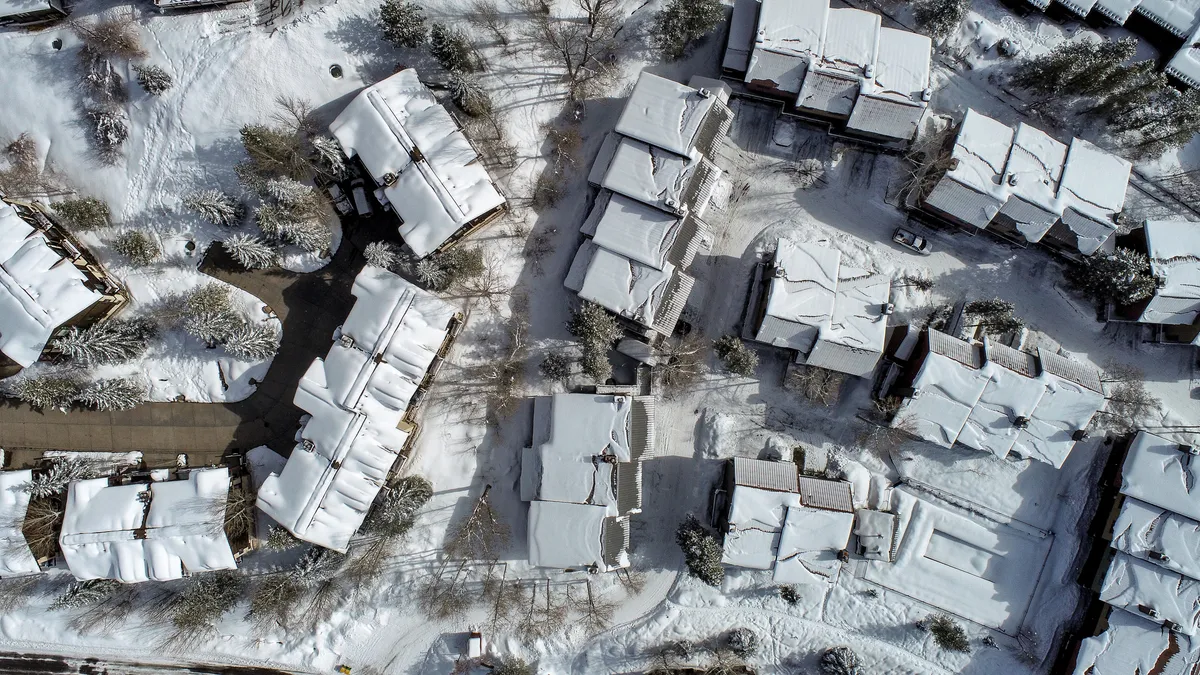 This exterior winter scene features snow-covered rooftops, tree-lined pathways, and neatly arranged buildings. The image evokes a serene winter mood and hints at the convenience of stepping outside for winter activities, with visible paths ideal for accessing the ski shuttle or enjoying a snowy walk.