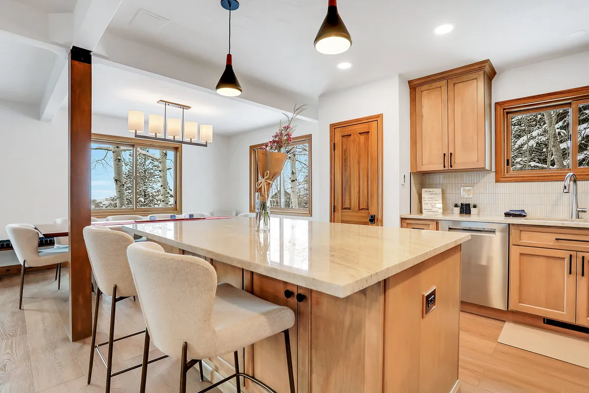 An open-concept kitchen and dining area with a large island countertop, stylish pendant lighting, and windows that overlook serene natural surroundings. A decorative vase with flowers adds a homey touch, while the ample seating fosters togetherness during mealtimes or celebrations.