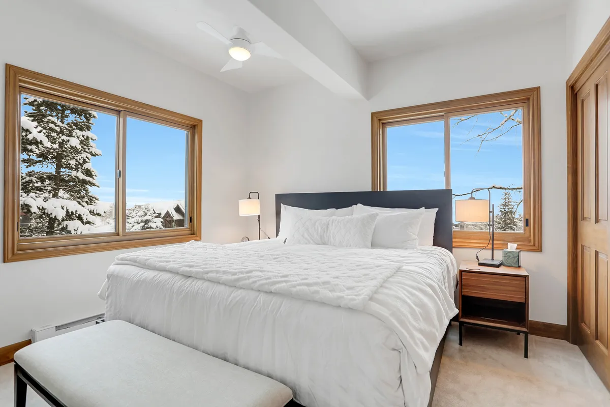 A serene bedroom with a bed featuring white bedding, large windows offering views of snow-covered trees, and a nightstand with a lamp. The natural light and winter landscape create an inviting, peaceful atmosphere perfect for relaxation after a day outdoors.