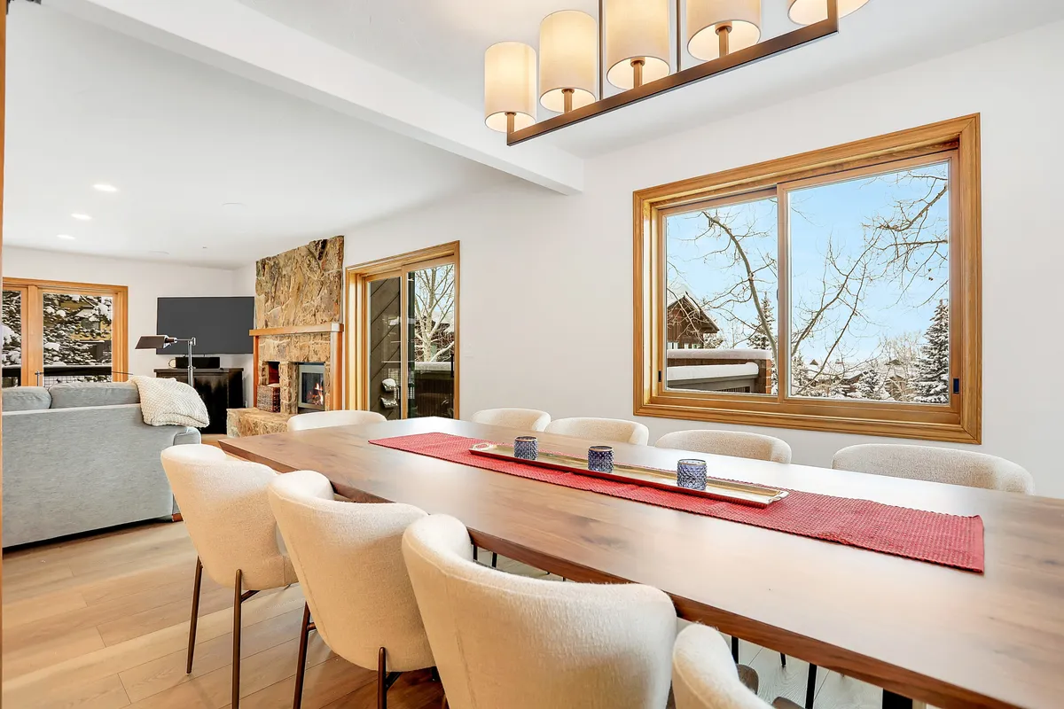 A long dining table that comfortably seats ten, positioned beneath a modern chandelier and flanked by large windows that let in abundant natural light. A cozy fireplace adds warmth and ambiance, making the space ideal for celebratory dinners, après-ski gatherings, or board games.