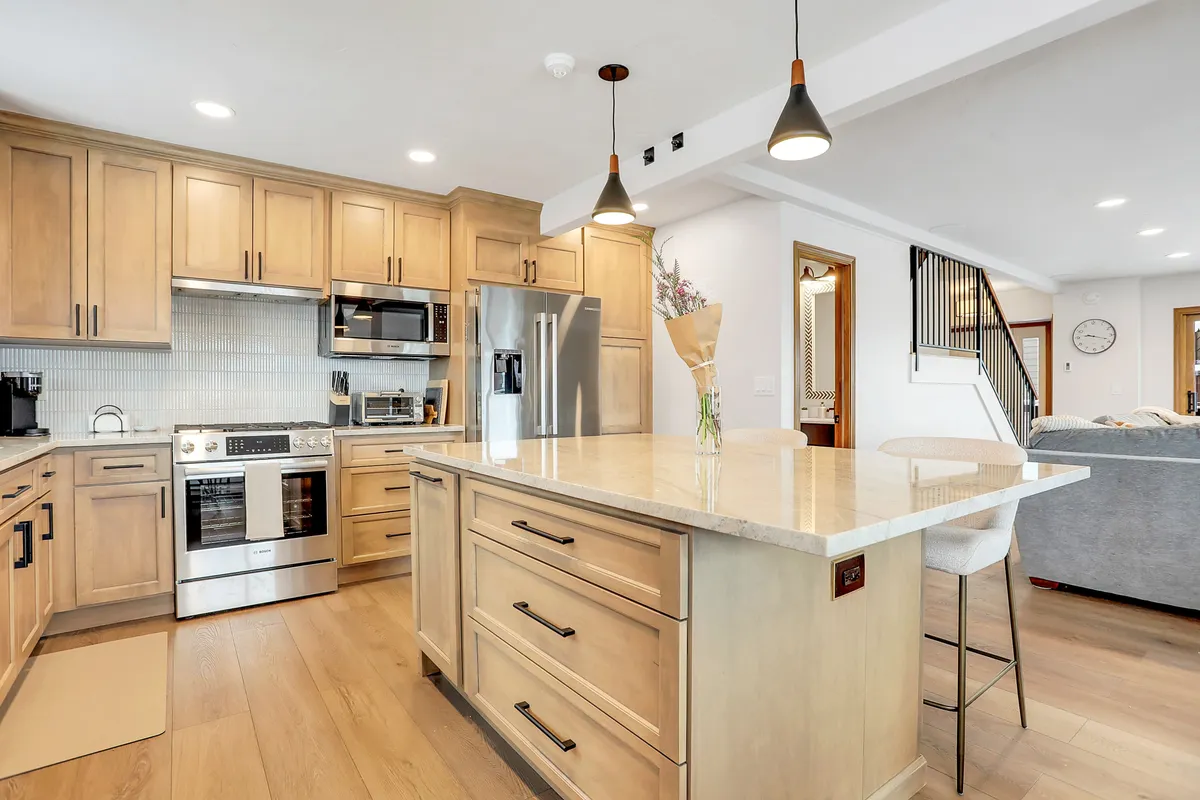 A bright, modern kitchen with a central island, sleek modern appliances—including an espresso machine—and warm wood cabinetry. The layout encourages both cooking and conversation, with the island doubling as a breakfast bar.