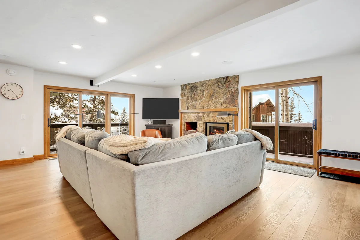A spacious living room featuring a large sectional sofa and a stone fireplace, with expansive windows offering beautiful outdoor views. The seating arrangement is ideal for group gatherings, family game nights, and cozy evenings together.