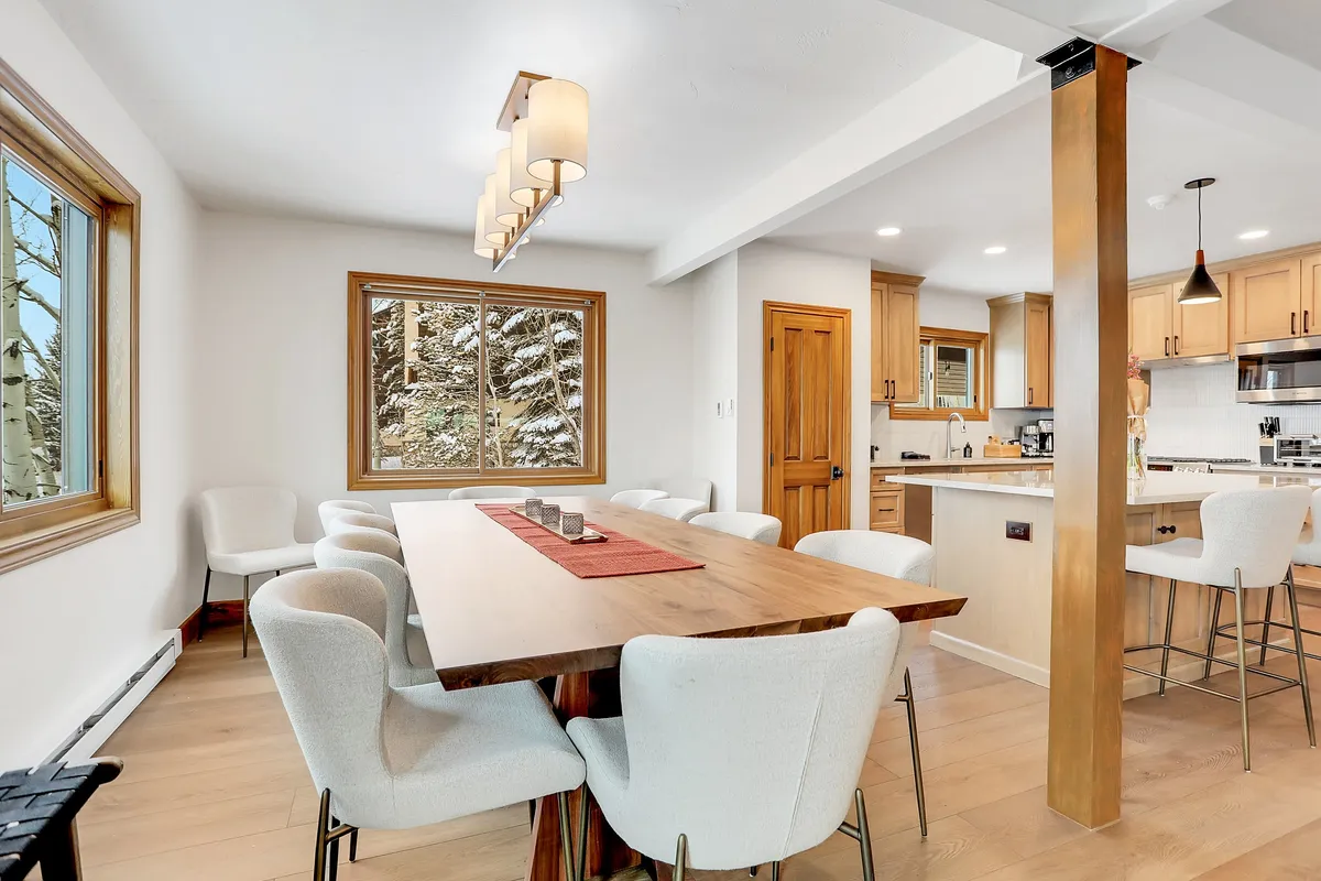 The image showcases a spacious and elegant dining area at Waterford 21, featuring a large wooden dining table, stylish light fixture, and a window framed by natural wood that offers inviting seasonal views. The scene evokes warmth and togetherness, ideal for family gatherings, dinner parties, or relaxing after a day on the slopes.