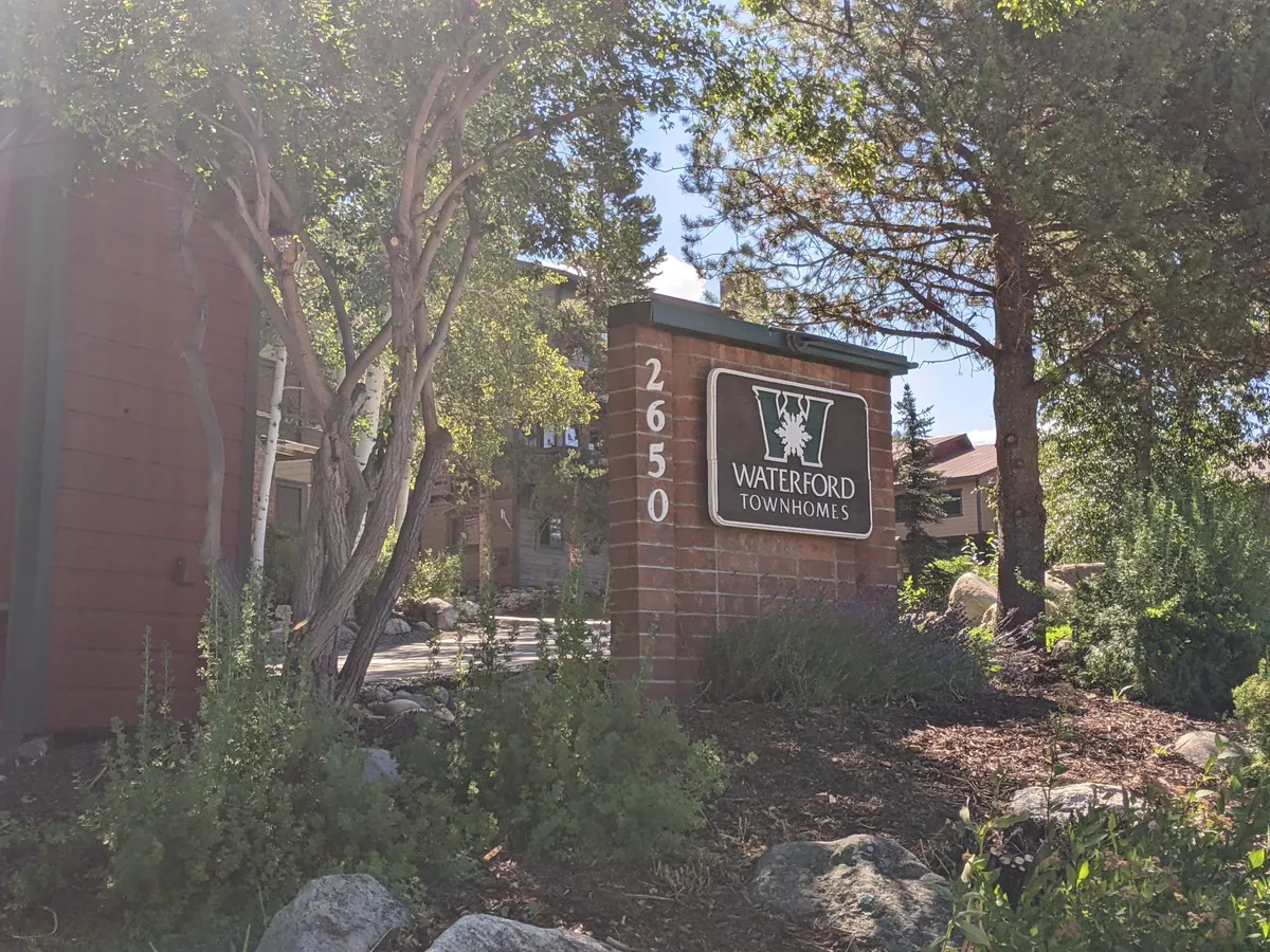 A welcoming entrance to the neighborhood featuring a townhome sign, manicured landscaped gardens, and tree-lined pathways. The inviting scene hints at easy access to walking and biking trails directly from the community.