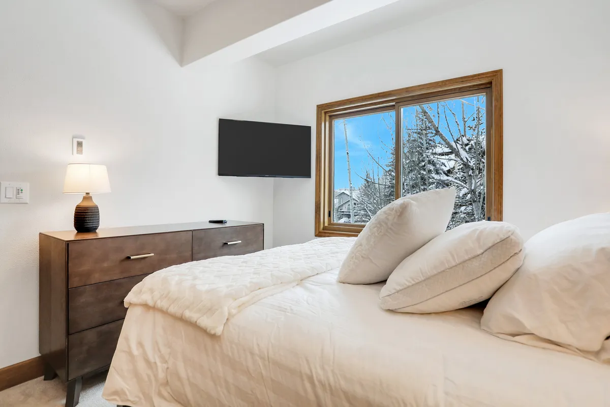 A tranquil bedroom featuring a plush bed, wooden dresser, and a large window that frames a snowy landscape outside. The room is styled for comfort, with soft bedding and peaceful lighting, while the view outside reminds guests of Steamboat's winter charm and the natural beauty just beyond their doorstep.
