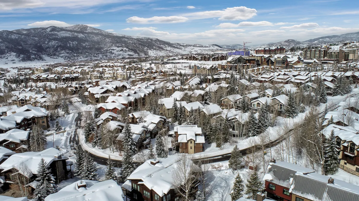 A serene exterior view featuring snow-capped rooftops, tall evergreens, a winding road, and distant mountains. The cluster of buildings suggests a welcoming neighborhood nestled in a winter landscape.